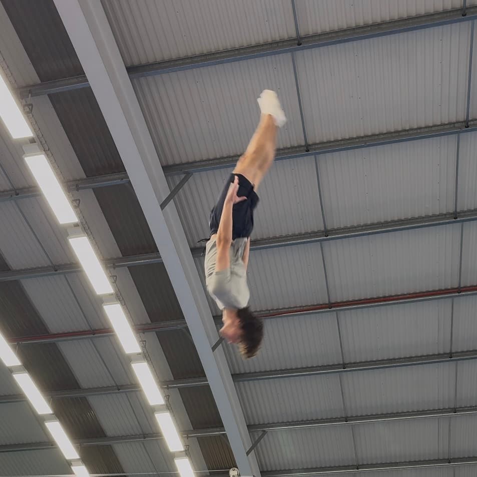 A trampolinist performing an inverted skill beneath a sports-hall roof
