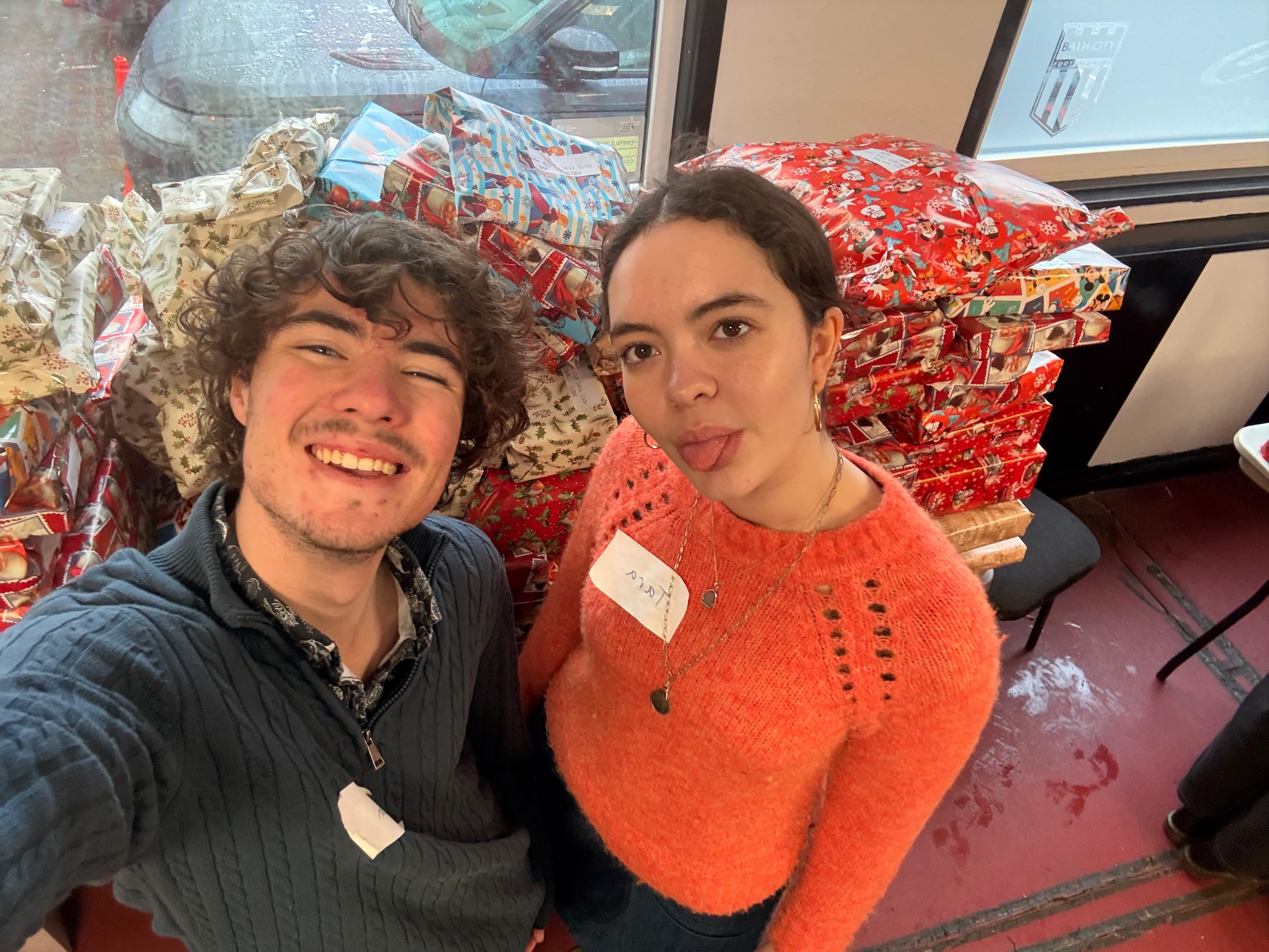 Preston Whiteman and his friend Tara-Mae Devine smiling in front of stacks of wrapped Christmas presents at the Wrapathon